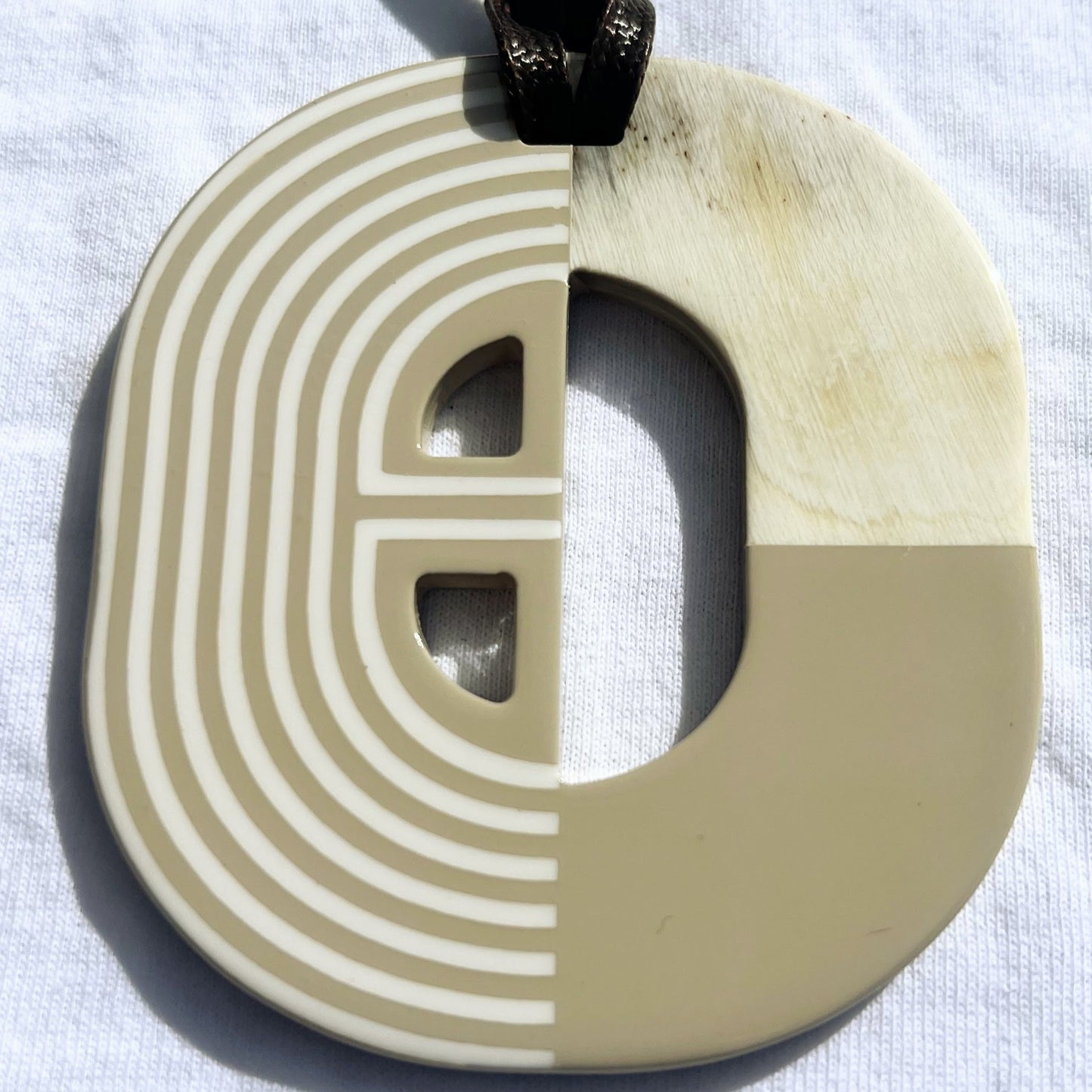 Pendentif en corne naturelle au design graphique bicolore rouge et ivoire. Sa forme semi-circulaire et ses lignes géométriques blanches contrastées apportent une touche d’élégance et de modernité. Il est suspendu à un cordon noir pour un style unique et audacieux.