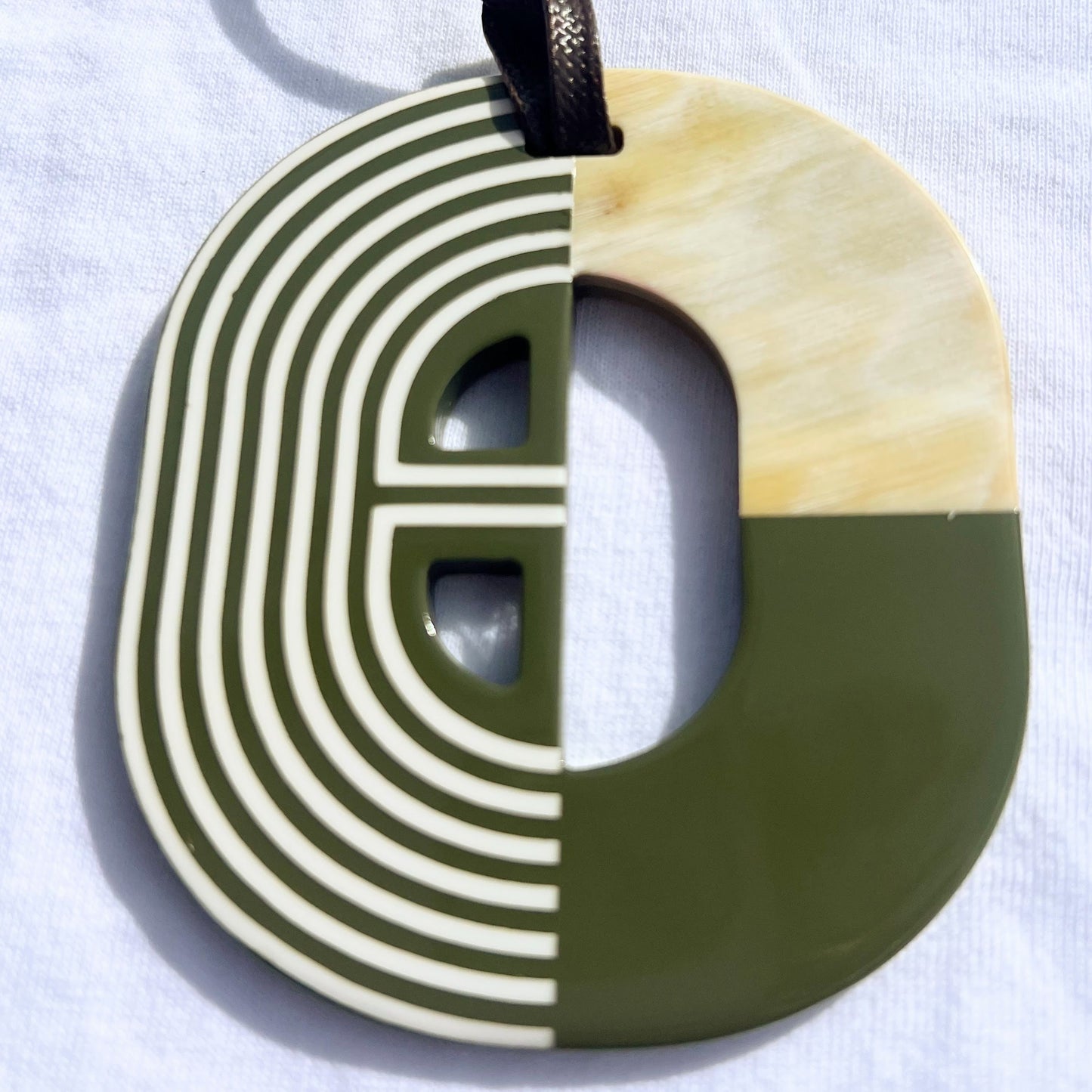 Pendentif en corne naturelle au design graphique bicolore rouge et ivoire. Sa forme semi-circulaire et ses lignes géométriques blanches contrastées apportent une touche d’élégance et de modernité. Il est suspendu à un cordon noir pour un style unique et audacieux.