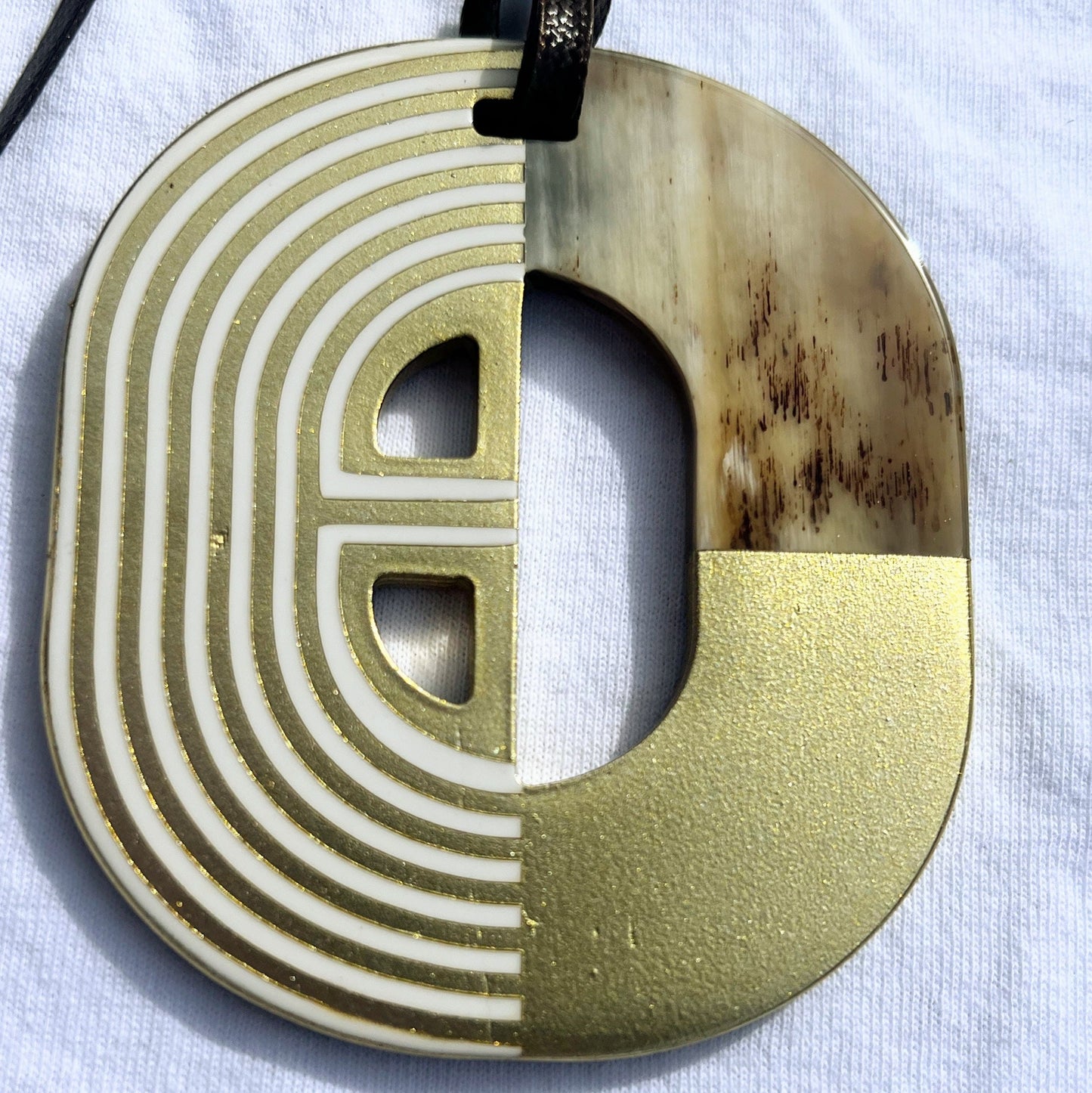 Pendentif en corne naturelle au design graphique bicolore rouge et ivoire. Sa forme semi-circulaire et ses lignes géométriques blanches contrastées apportent une touche d’élégance et de modernité. Il est suspendu à un cordon noir pour un style unique et audacieux.