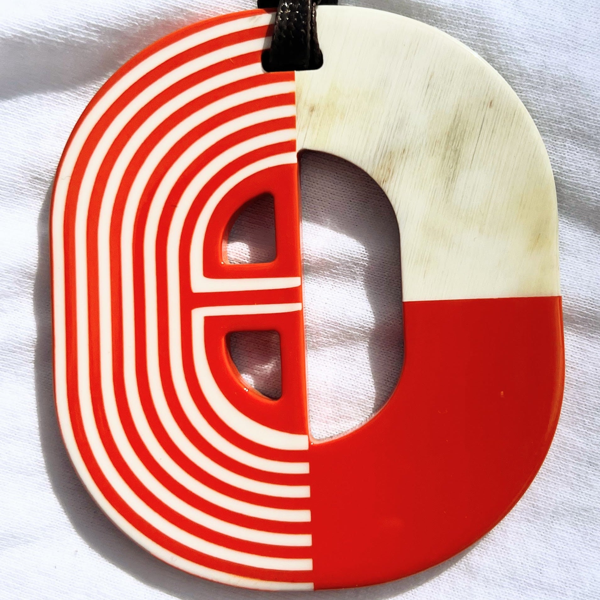  Pendentif en corne naturelle au design graphique bicolore rouge et ivoire. Sa forme semi-circulaire et ses lignes géométriques blanches contrastées apportent une touche d’élégance et de modernité. Il est suspendu à un cordon noir pour un style unique et audacieux.

