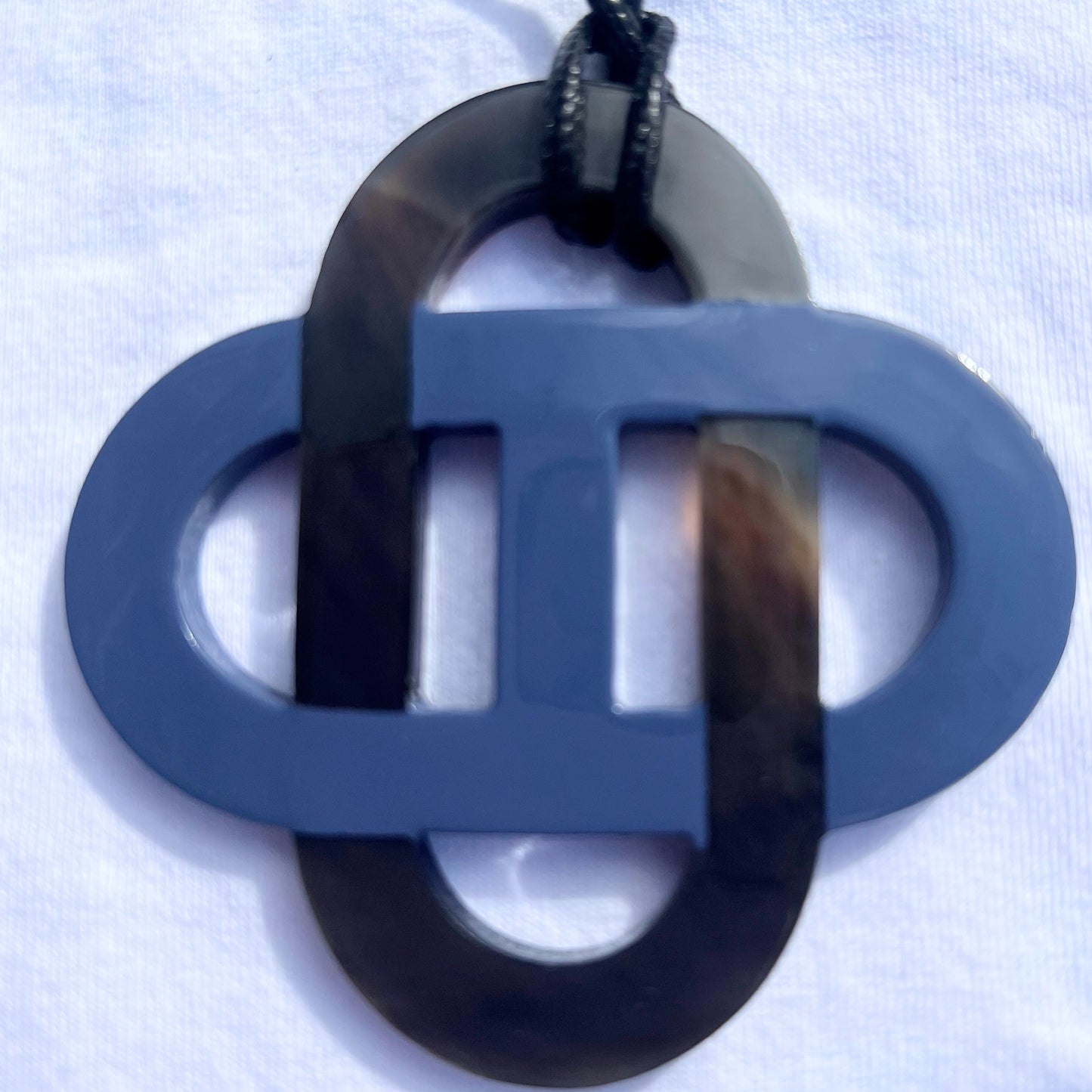 Pendentif en corne naturelle noir et beige au design symétrique inspiré des motifs entrelacés. Son jeu de contrastes entre la corne brute et la partie noire lui confère un style raffiné et intemporel. Il est monté sur un cordon noir ajustable.