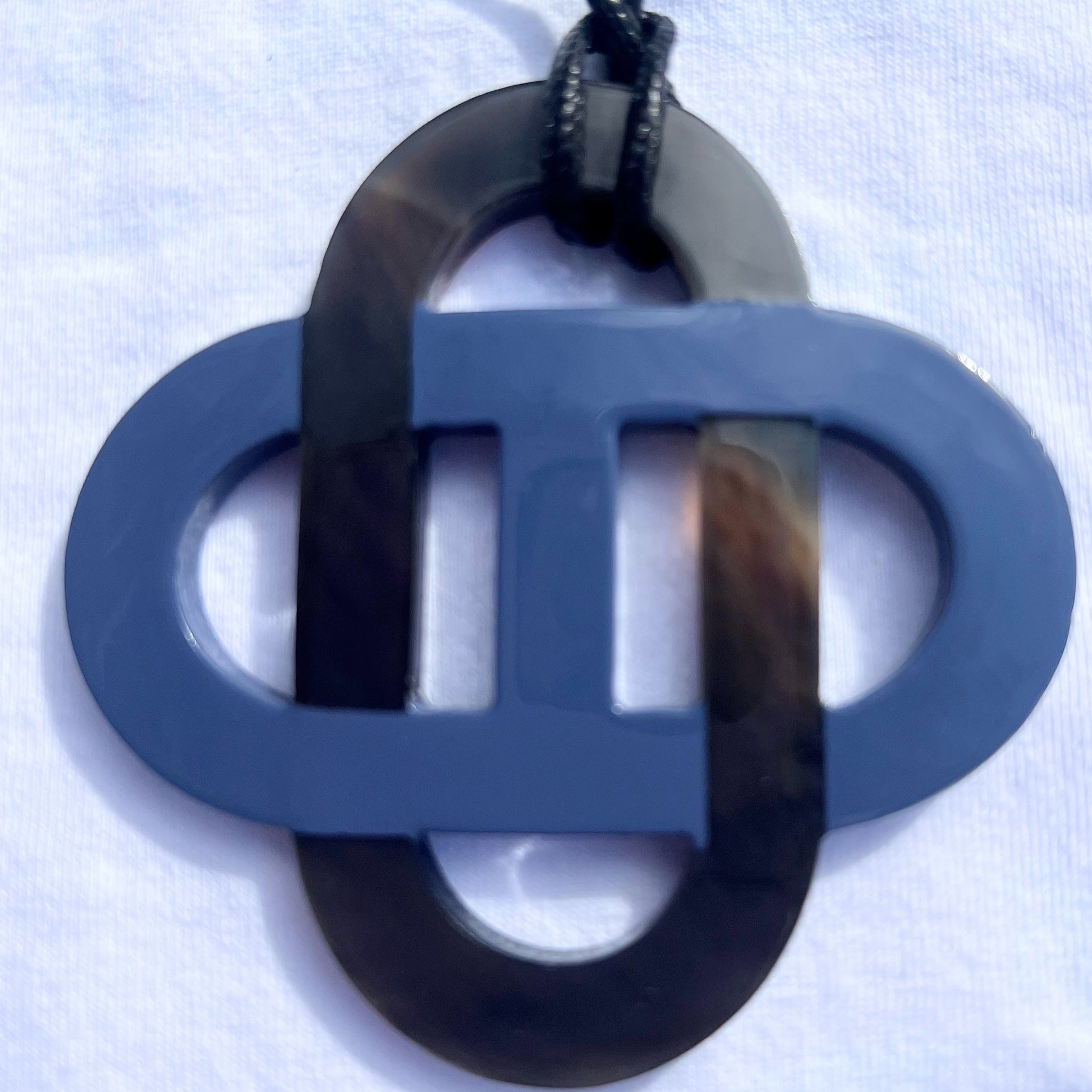 Pendentif en corne naturelle noir et beige au design symétrique inspiré des motifs entrelacés. Son jeu de contrastes entre la corne brute et la partie noire lui confère un style raffiné et intemporel. Il est monté sur un cordon noir ajustable.