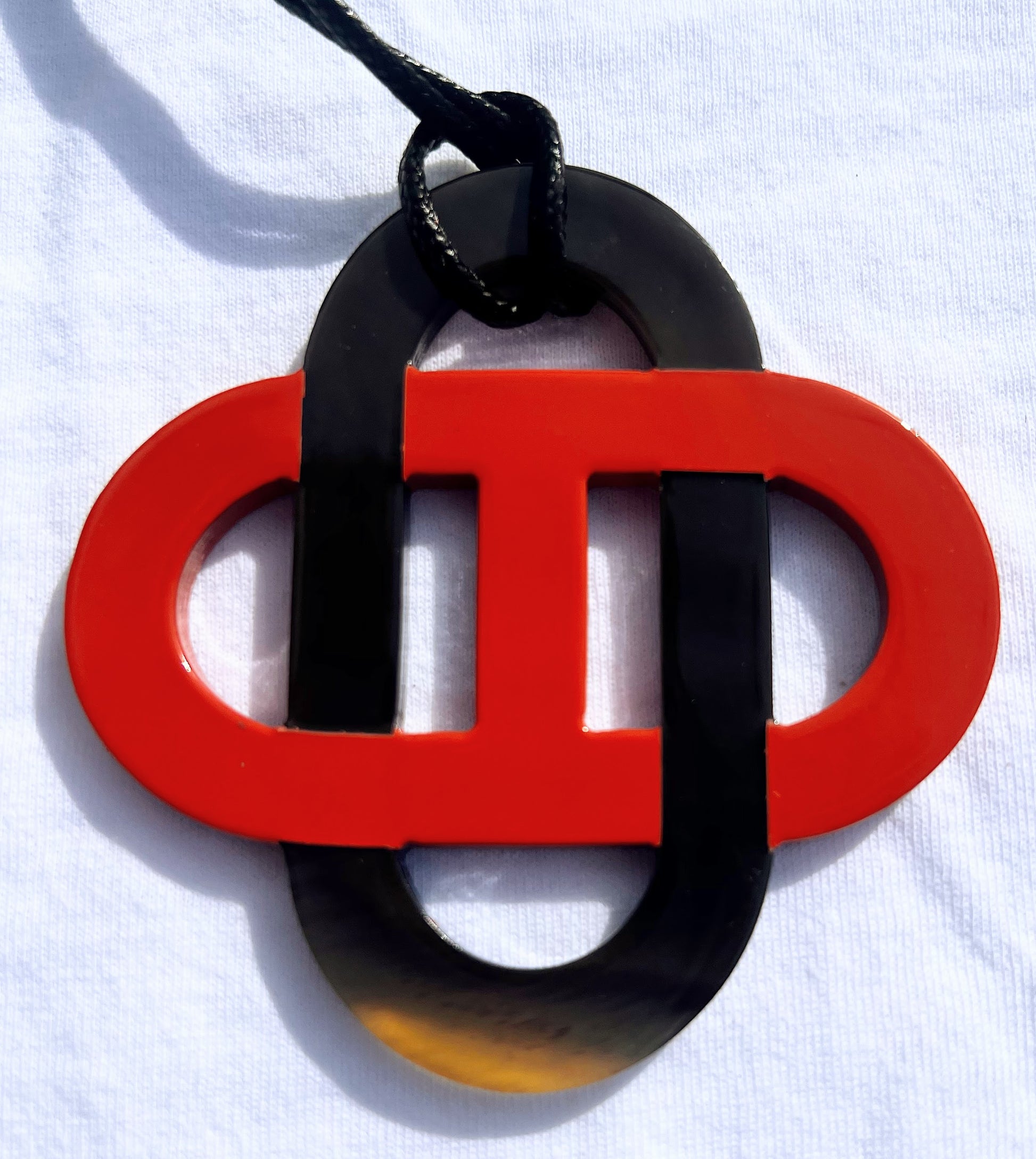 Pendentif en corne naturelle noir et beige au design symétrique inspiré des motifs entrelacés. Son jeu de contrastes entre la corne brute et la partie noire lui confère un style raffiné et intemporel. Il est monté sur un cordon noir ajustable.