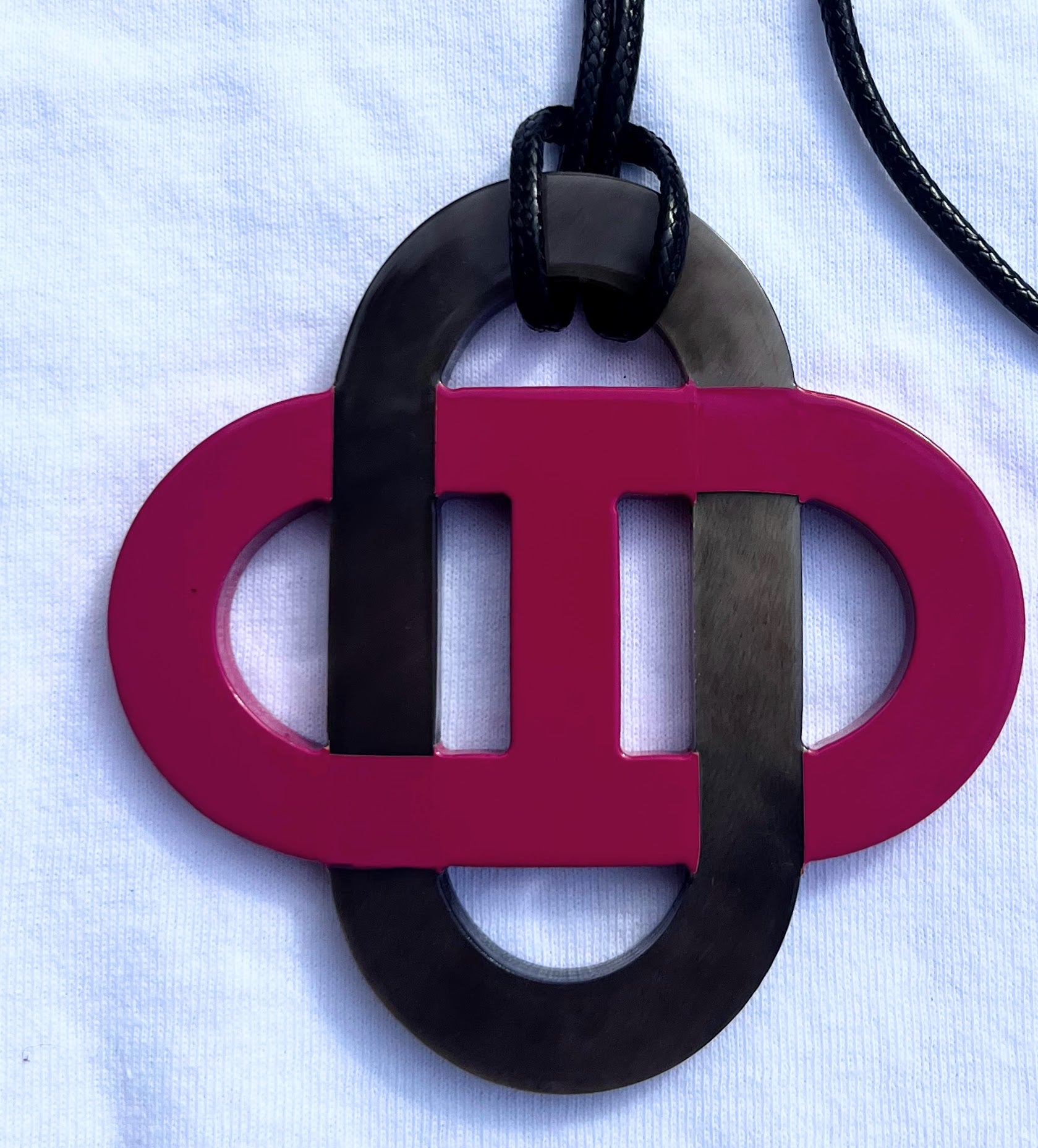 Pendentif en corne naturelle noir et beige au design symétrique inspiré des motifs entrelacés. Son jeu de contrastes entre la corne brute et la partie noire lui confère un style raffiné et intemporel. Il est monté sur un cordon noir ajustable.