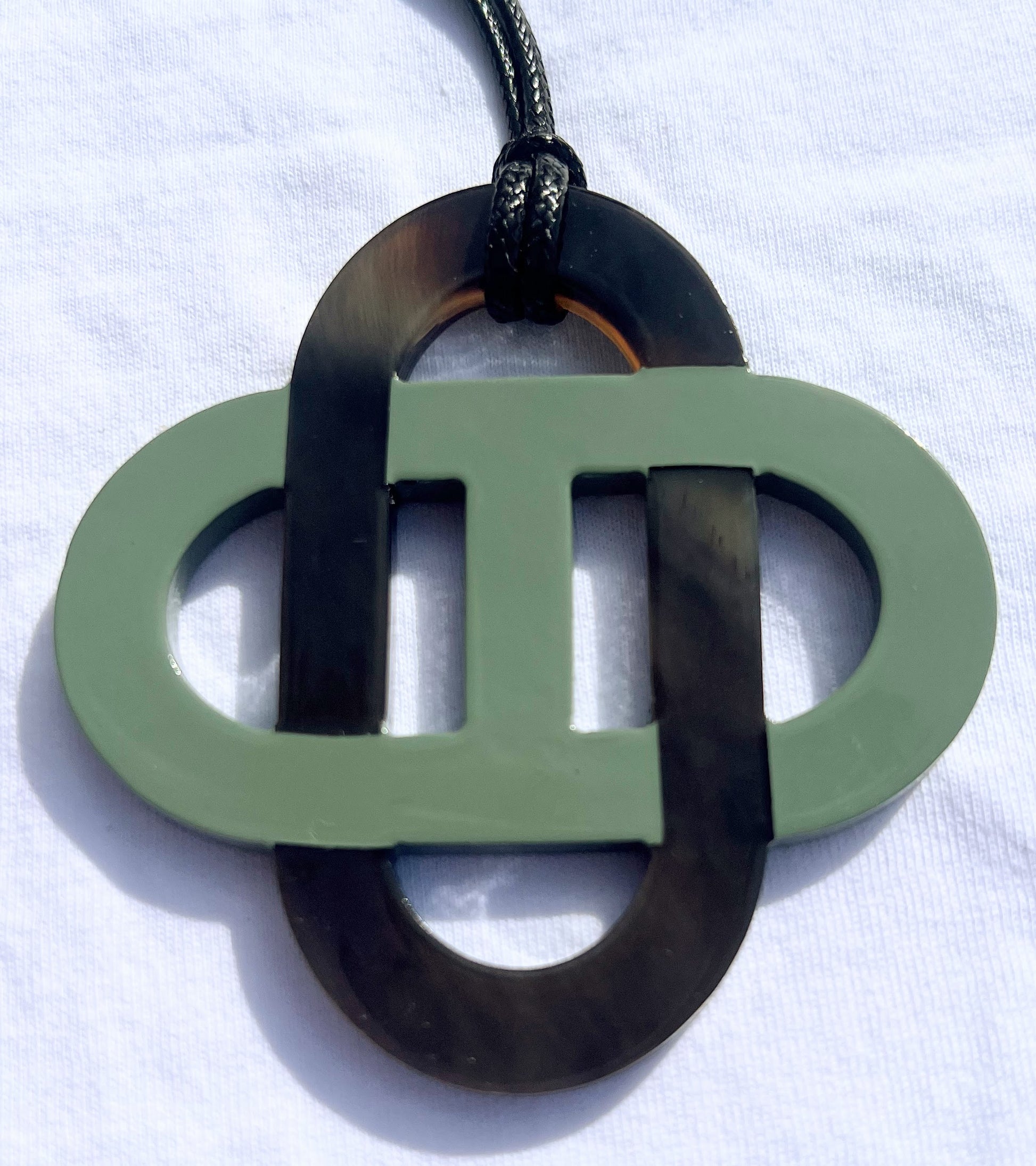Pendentif en corne naturelle noir et beige au design symétrique inspiré des motifs entrelacés. Son jeu de contrastes entre la corne brute et la partie noire lui confère un style raffiné et intemporel. Il est monté sur un cordon noir ajustable.