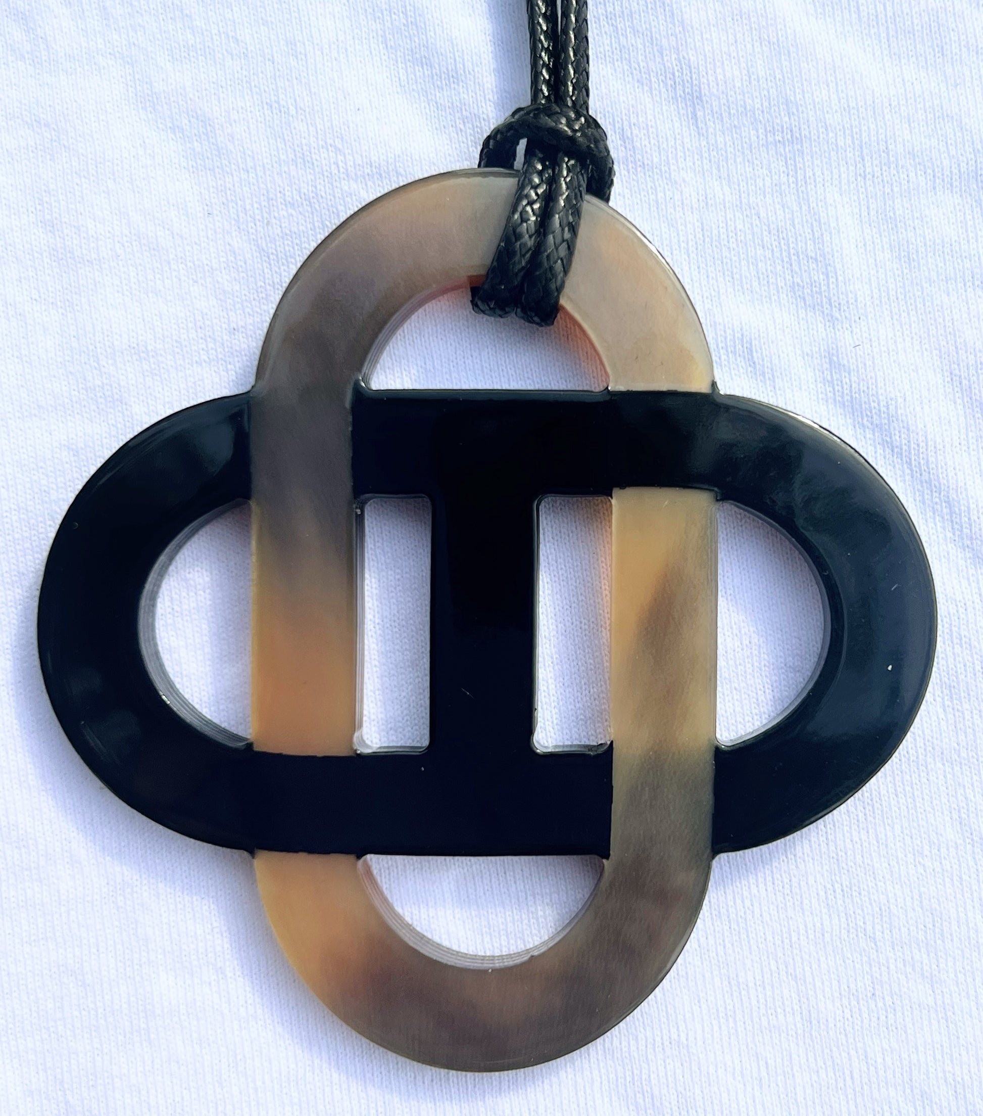 Pendentif en corne naturelle noir et beige au design symétrique inspiré des motifs entrelacés. Son jeu de contrastes entre la corne brute et la partie noire lui confère un style raffiné et intemporel. Il est monté sur un cordon noir ajustable.